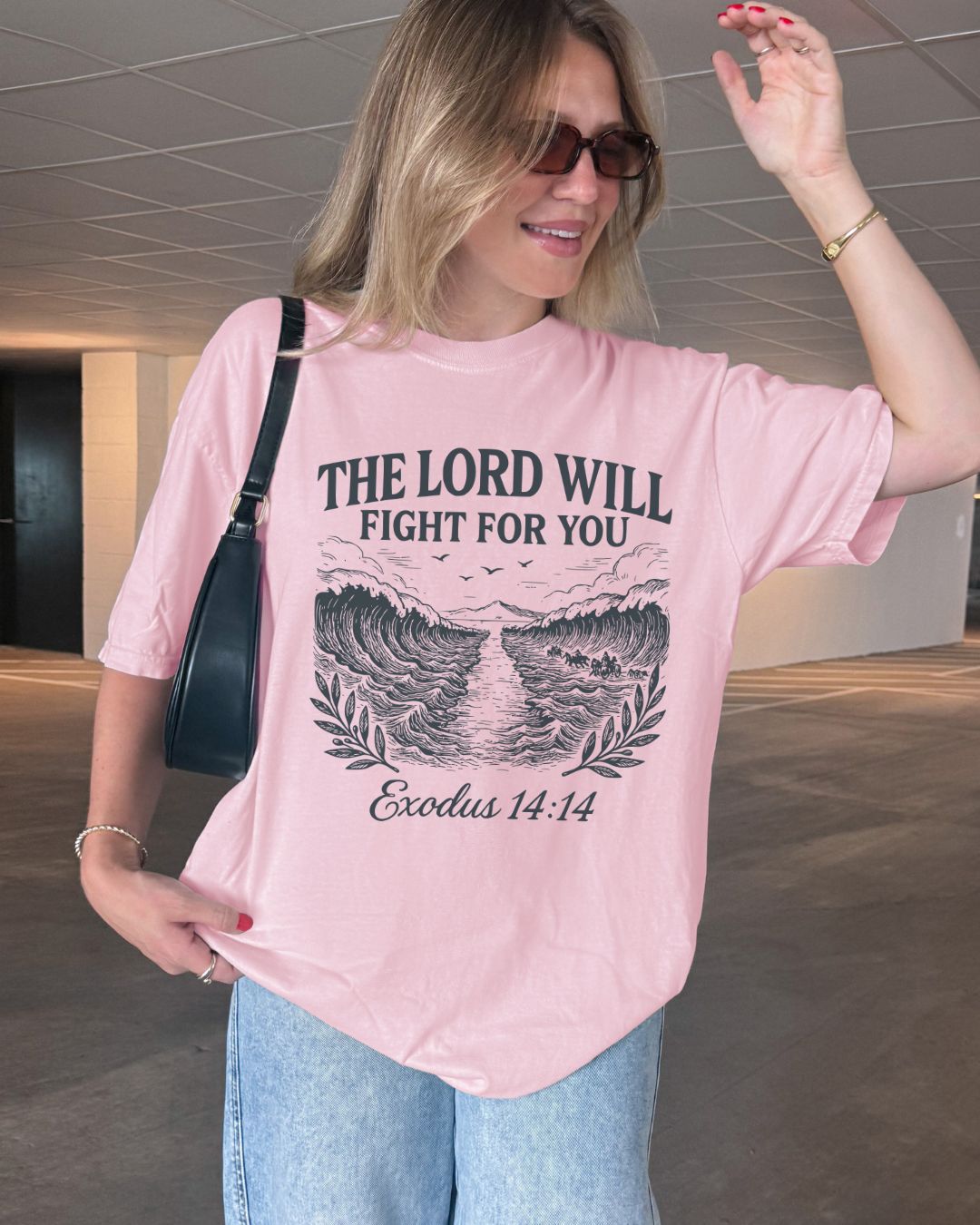 A Christian Woman wearing a pink t-shirt with Exodus 14:14, The Lord Will Fight For You, sitting on a white background.