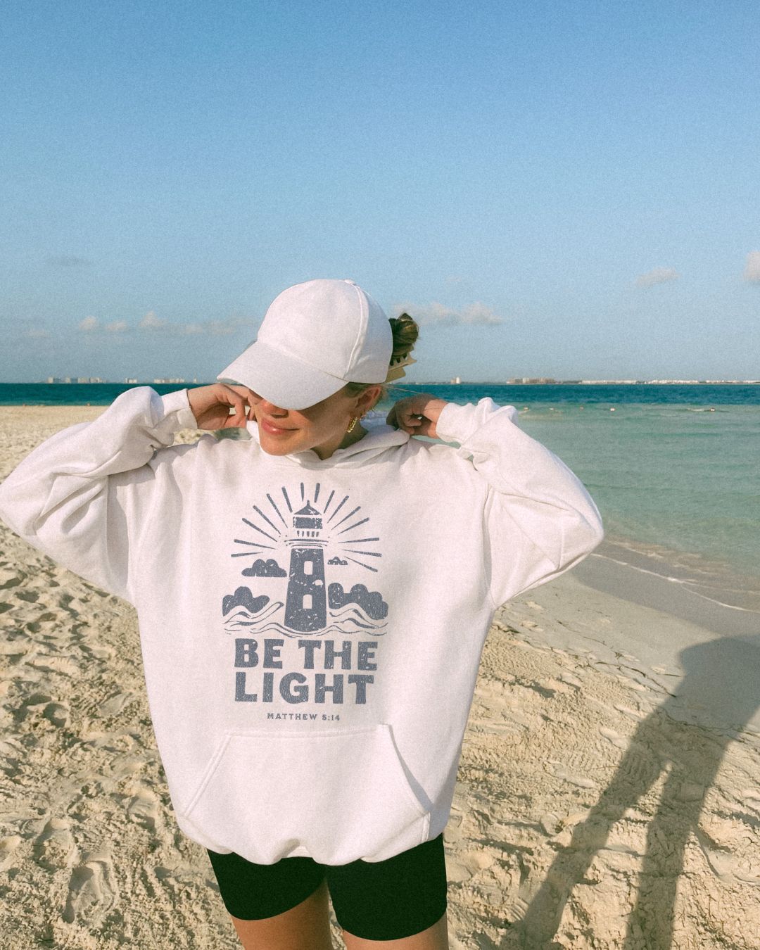 Woman wearing white Be The Light Christian hoodie on beach