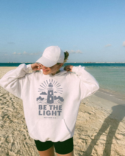 Woman wearing white Be The Light Christian hoodie on beach