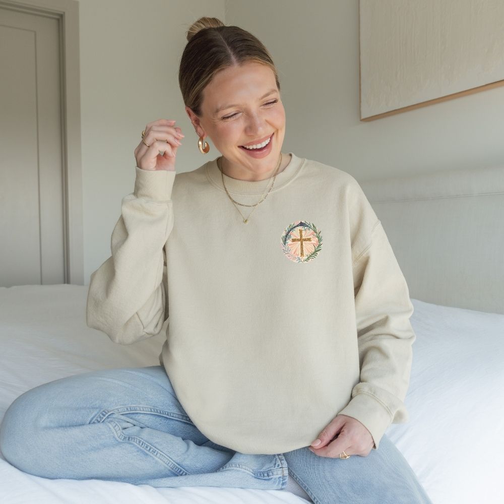 Woman wearing a beige sweatshirt with a logo, sitting on a bed in a room.