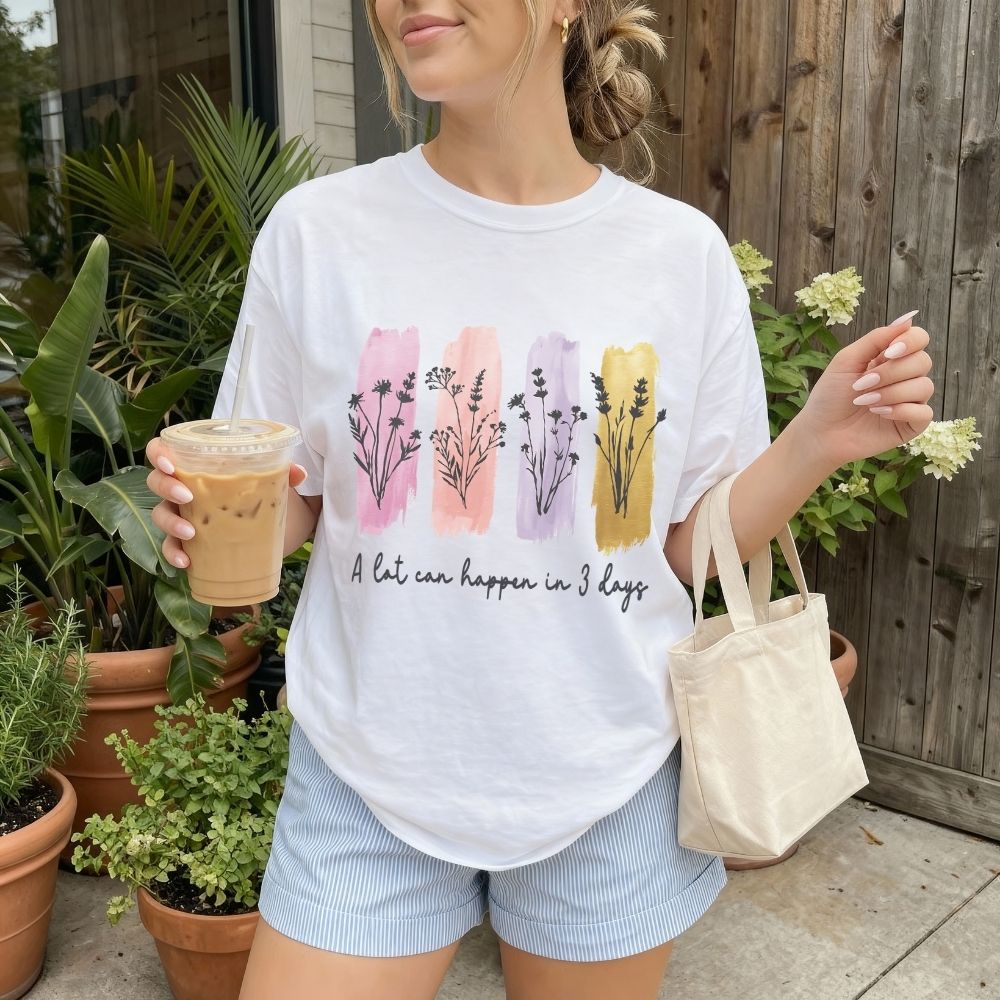 Woman wearing a white t-shirt with floral designs and text, holding a drink and a tote bag outdoors.