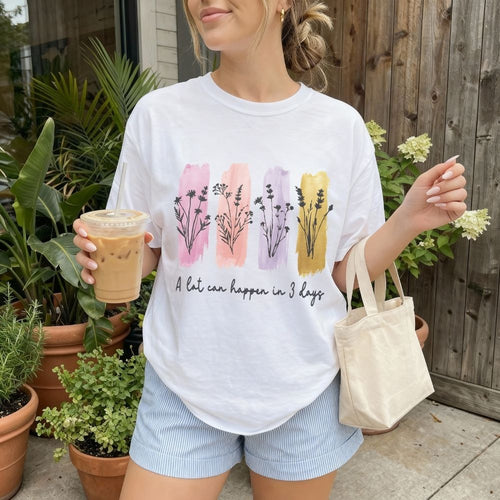 Woman wearing a white t-shirt with floral designs and text, holding a drink and a tote bag outdoors.