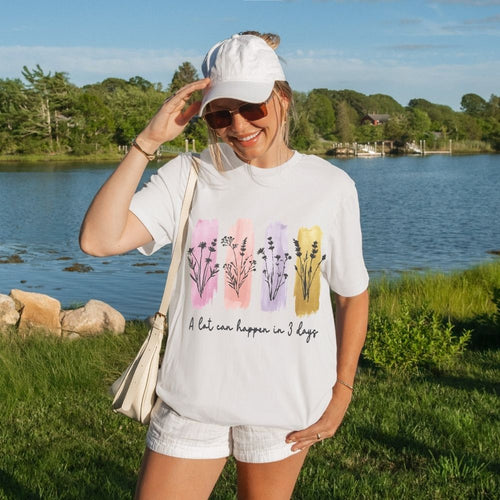 Woman wearing a white t-shirt with floral designs and text, standing by a lake.