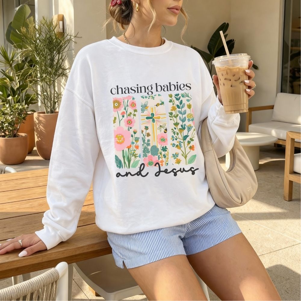 Woman wearing a white sweatshirt with floral design and text, holding a drink outdoors.