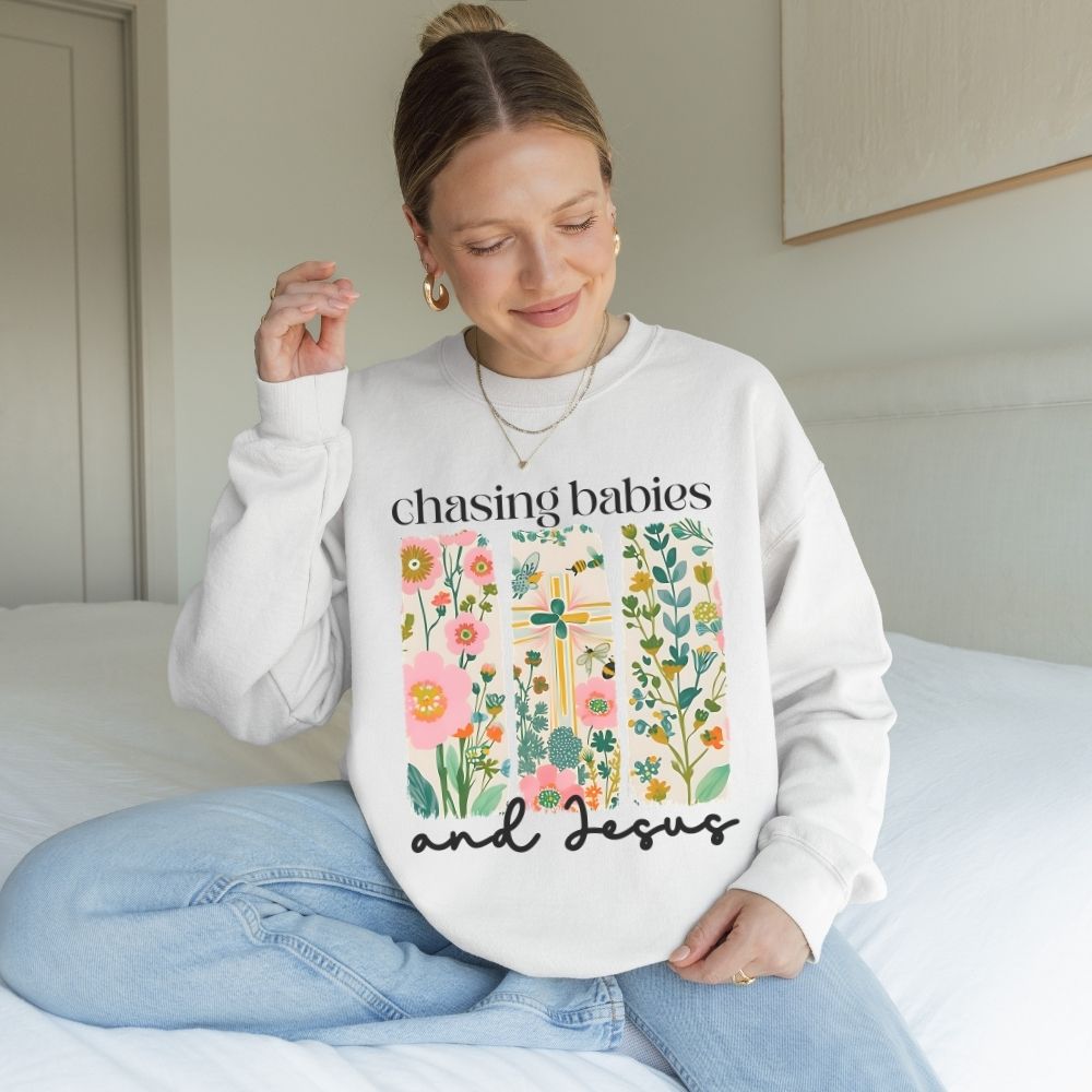 Woman wearing a white sweatshirt with floral design and text, sitting on a bed.
