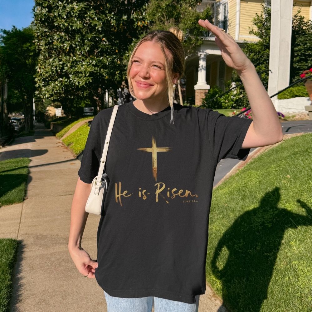 Person wearing a black t-shirt with 'He is Risen' text and a cross, standing outdoors.