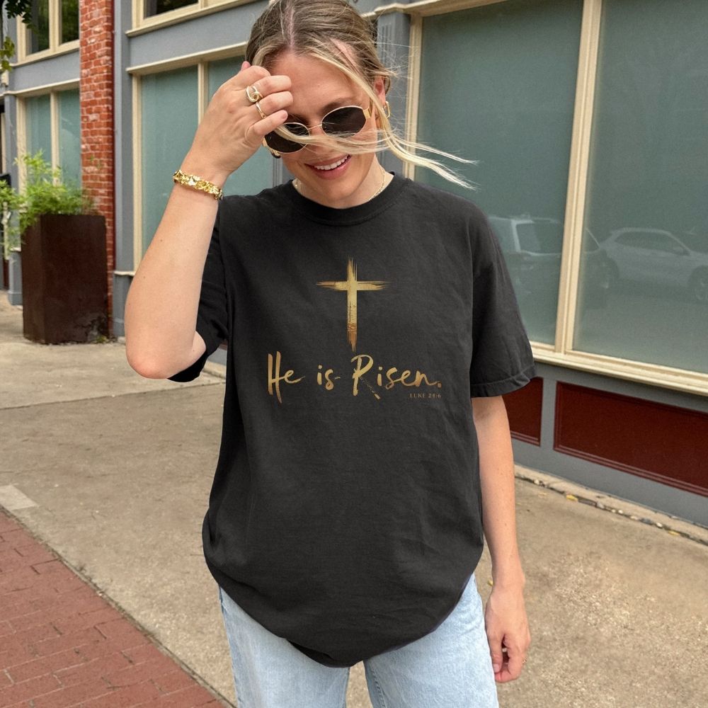Person wearing a black t-shirt with 'He is Risen' text and a cross symbol outdoors.