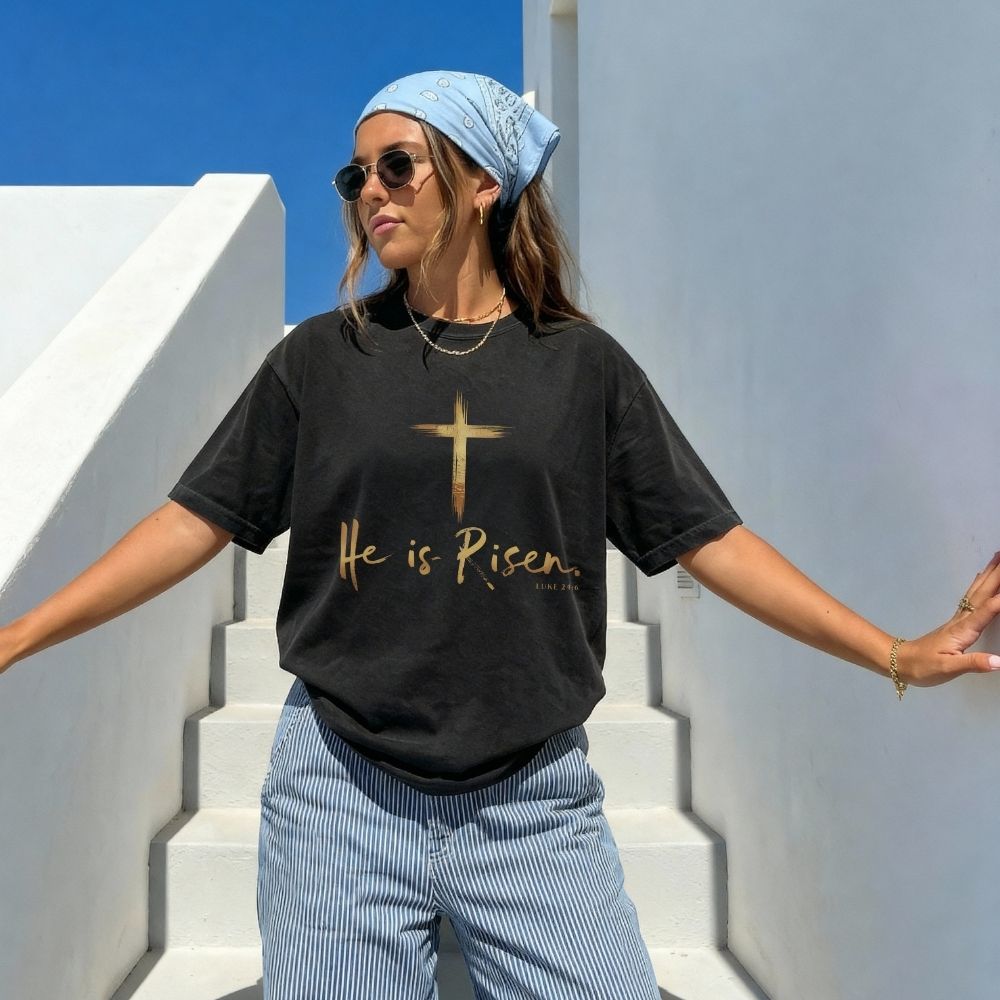 Woman wearing a black t-shirt with 'He is Risen' text and a cross symbol, standing on white steps with blue sky.
