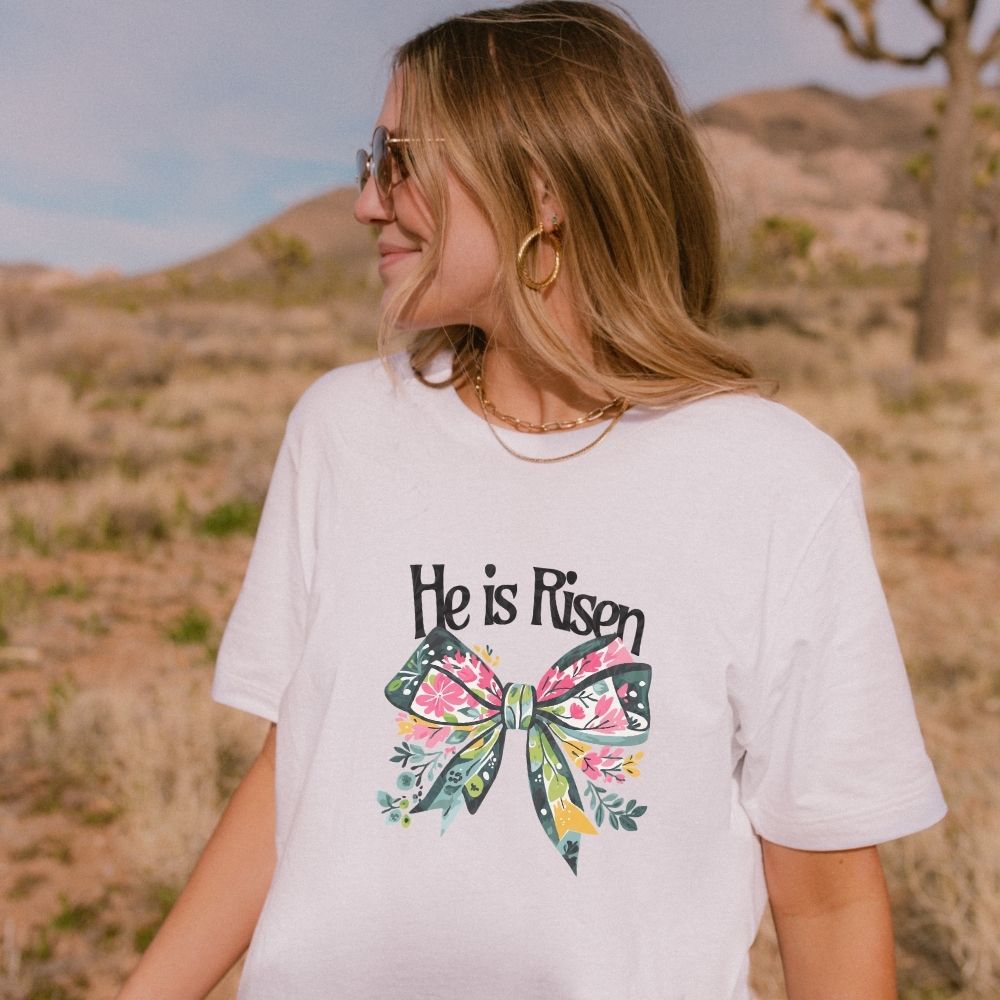 Woman wearing a white t-shirt with 'He is Risen' and a floral bow design in a desert setting.