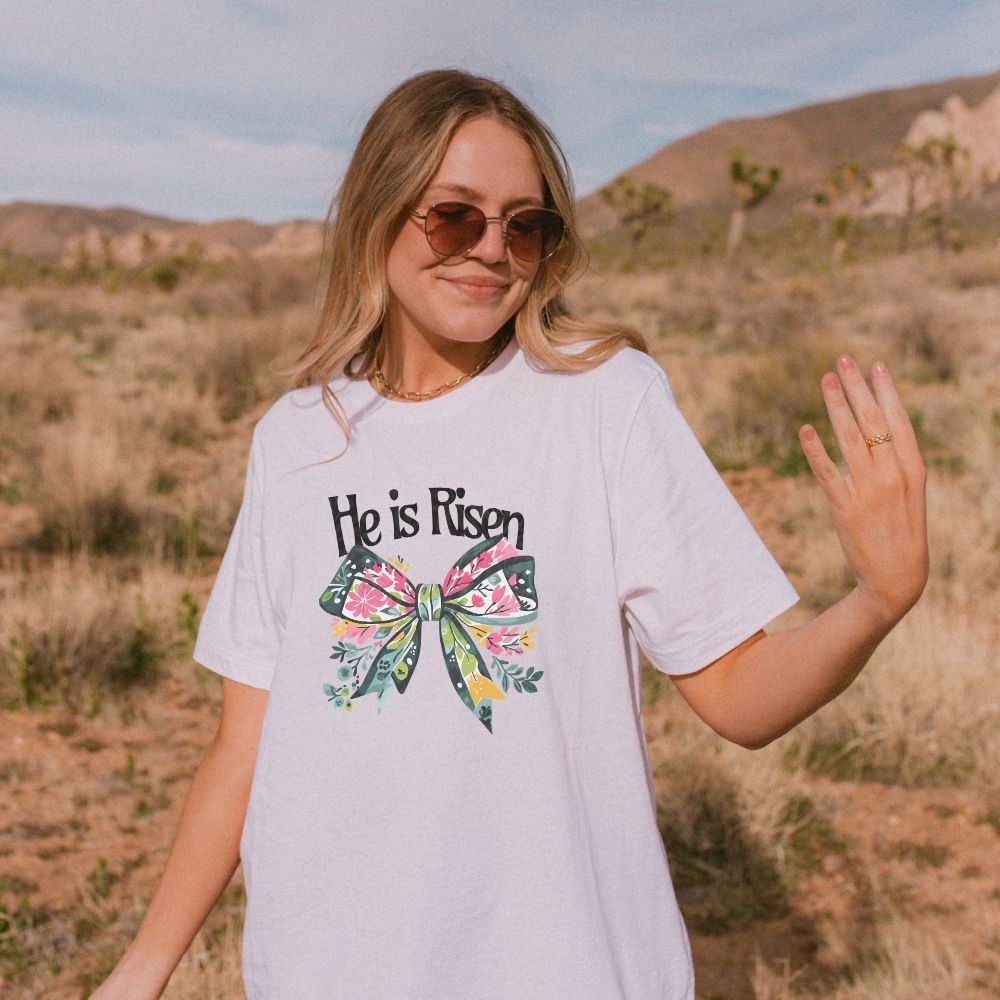 Woman wearing a t-shirt with 'He is Risen' and a floral bow design in a desert setting