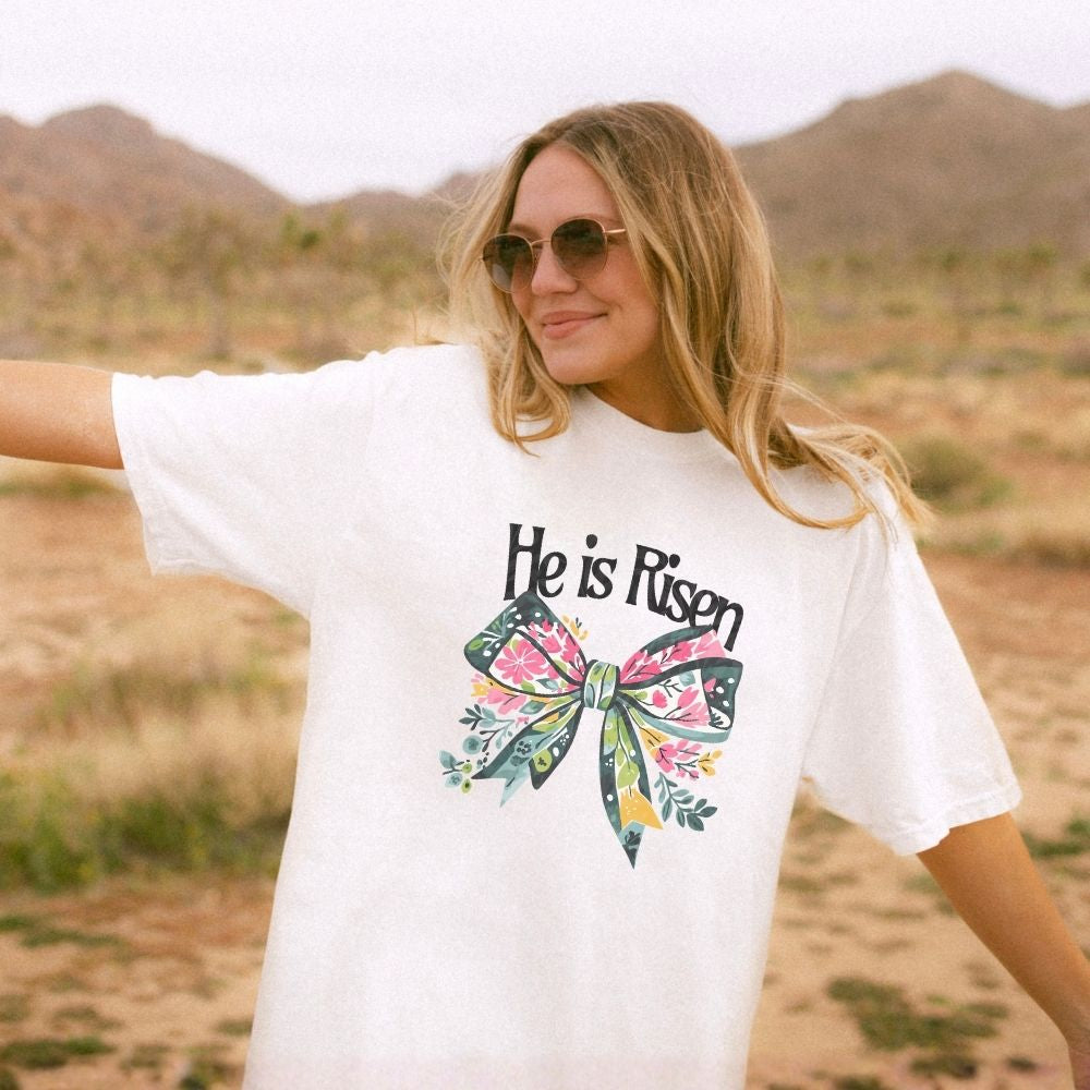 Woman wearing a white t-shirt with 'He is Risen' design in a desert setting