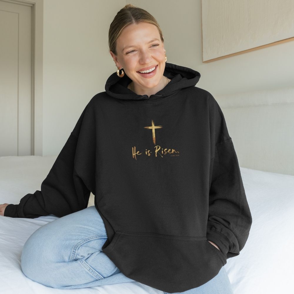 Person wearing a black hoodie with 'He is Risen' text and a cross symbol, sitting on a bed.