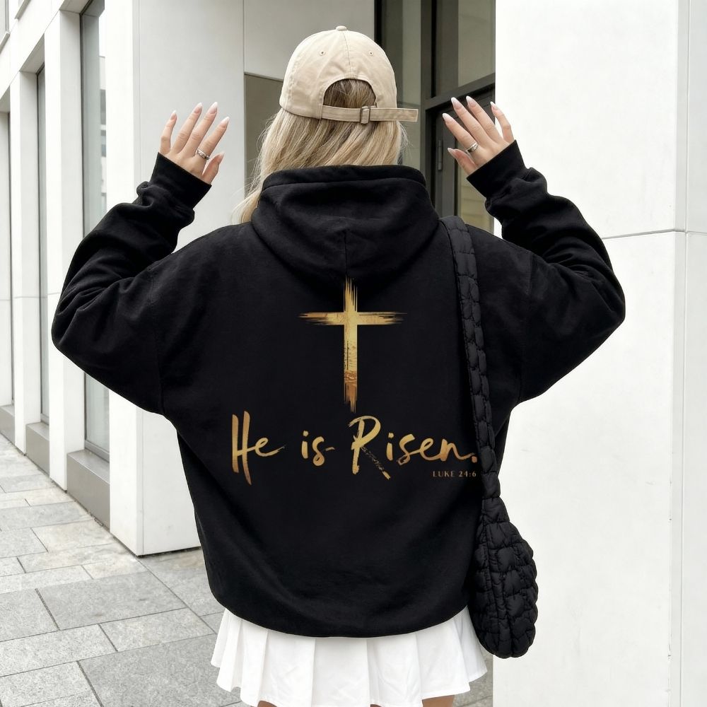 Person wearing a black hoodie with 'He is Risen' text and cross design on the back, standing in an urban setting.