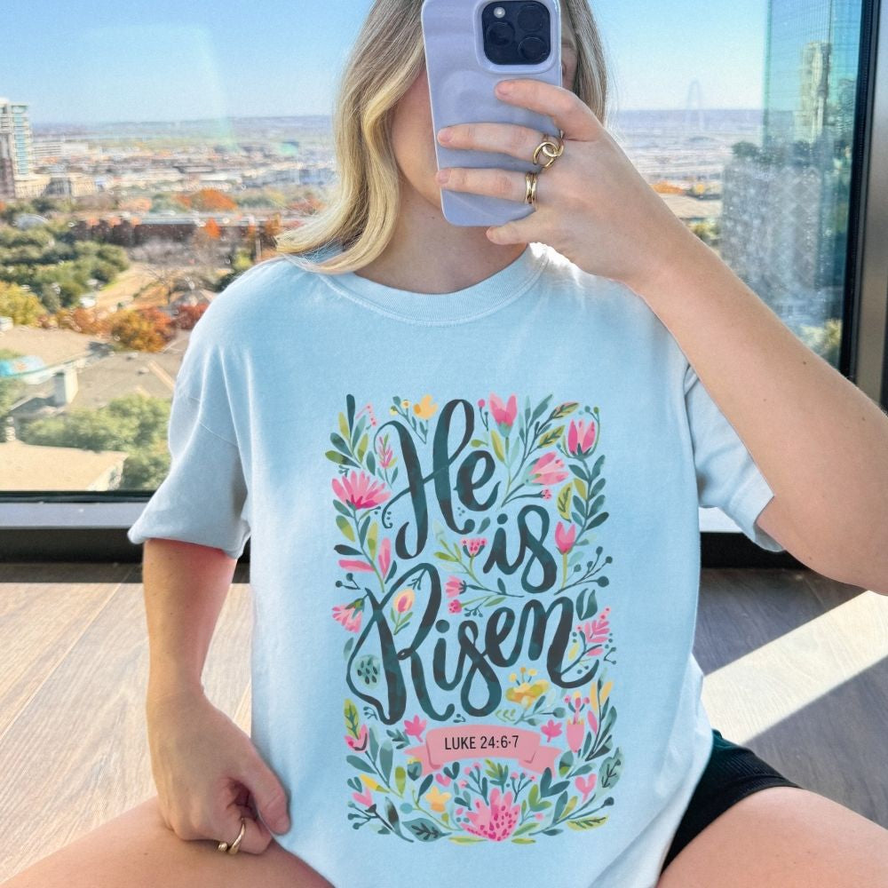 Person wearing a light blue t-shirt with 'He is Risen' design, taking a selfie in a high-rise setting.