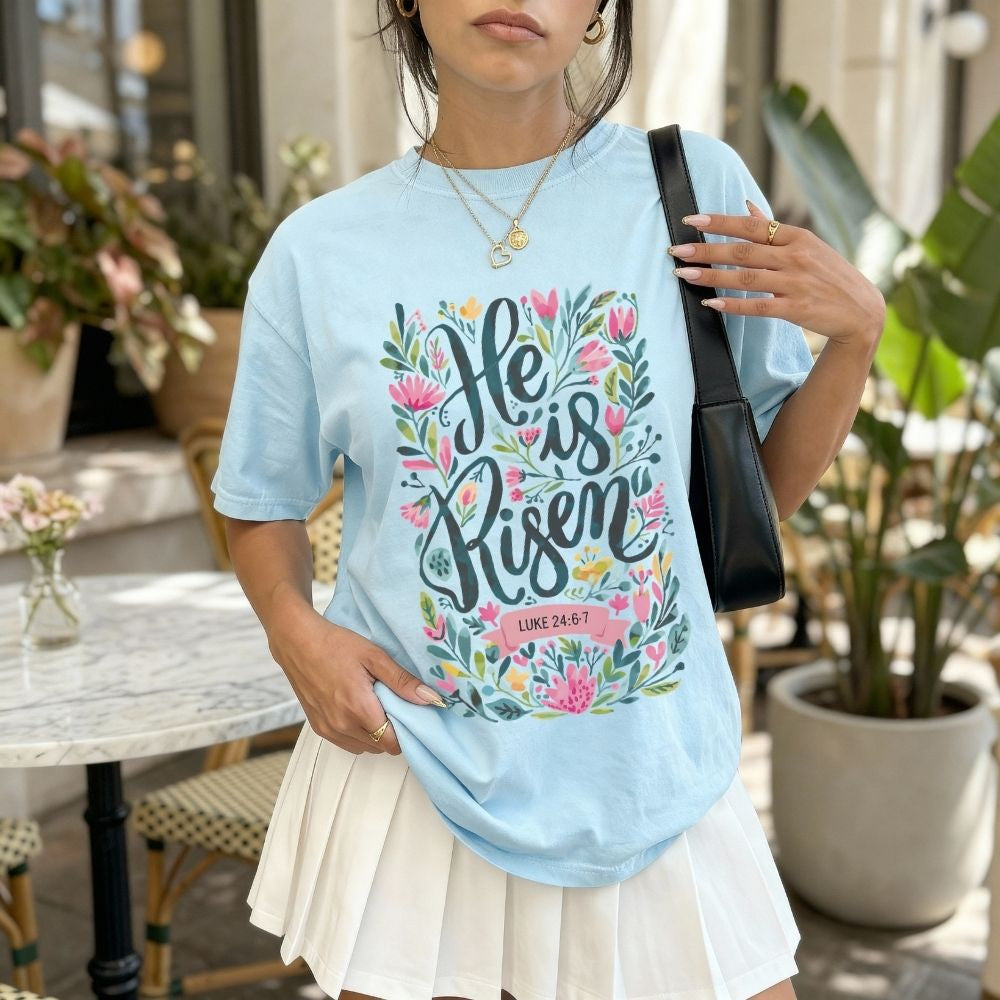 Woman wearing a light blue t-shirt with 'He is Risen' design outdoors.