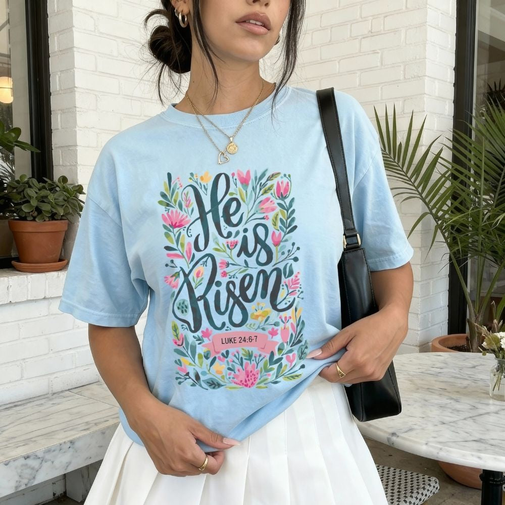 Woman wearing a light blue t-shirt with 'He is Risen' design outdoors.