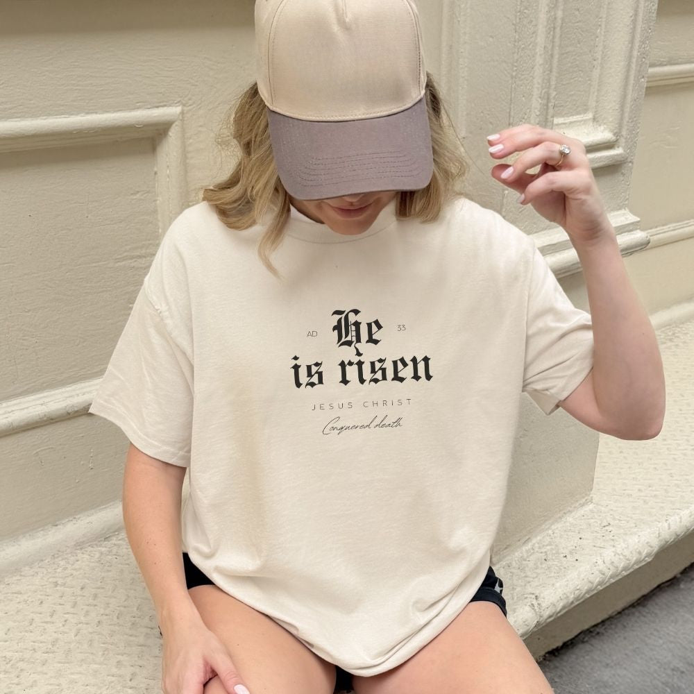 Person wearing a white t-shirt with 'He is risen' text sitting on stairs.