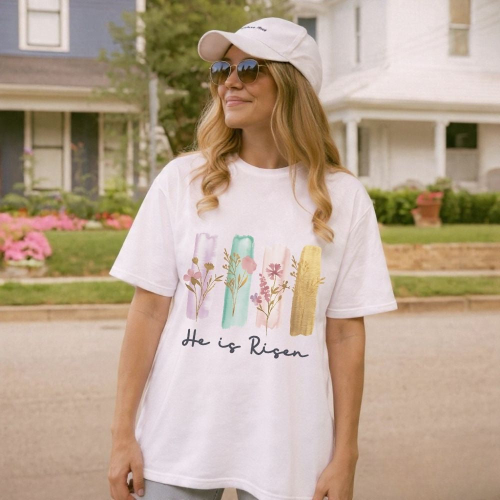 Woman wearing a white t-shirt with 'He is Risen' text and floral design outdoors.