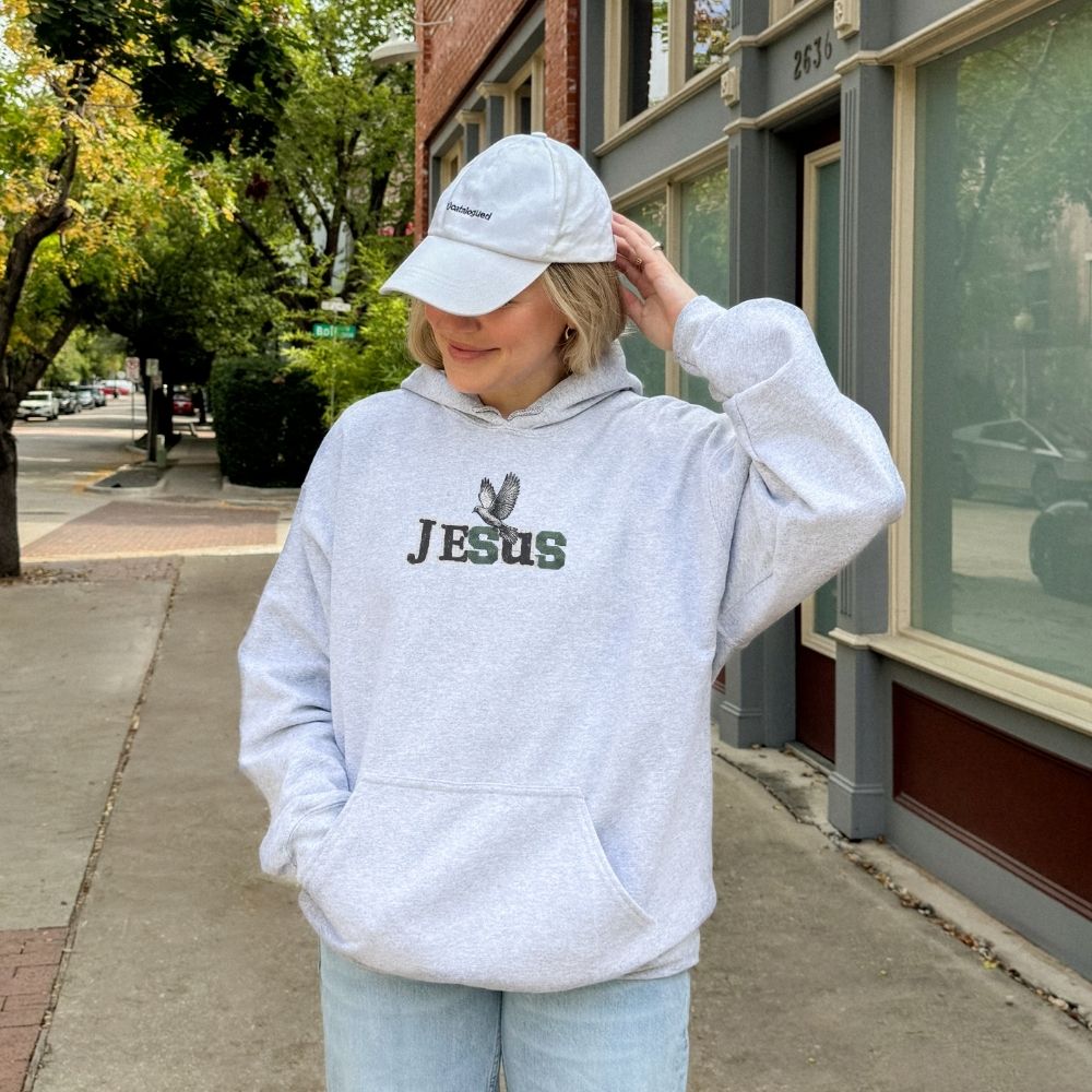Person wearing a gray hoodie with 'JESUS' printed on it, standing on a sidewalk.