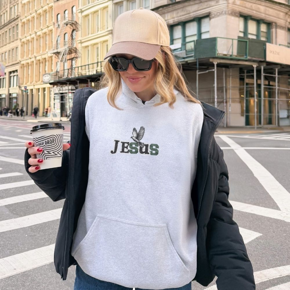 Person wearing a hoodie with 'Jesus' printed on it, standing on a city street.