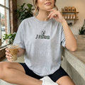 Person wearing a gray t-shirt with 'JESUS' printed on it, holding a drink in a casual setting.