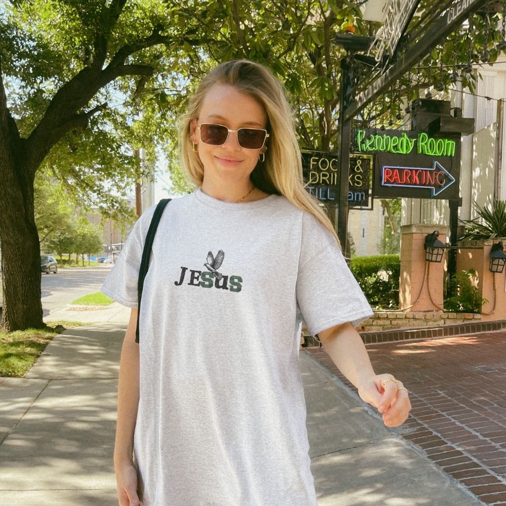 Person wearing a white t-shirt with 'Jesus' printed on it, standing on a sidewalk.