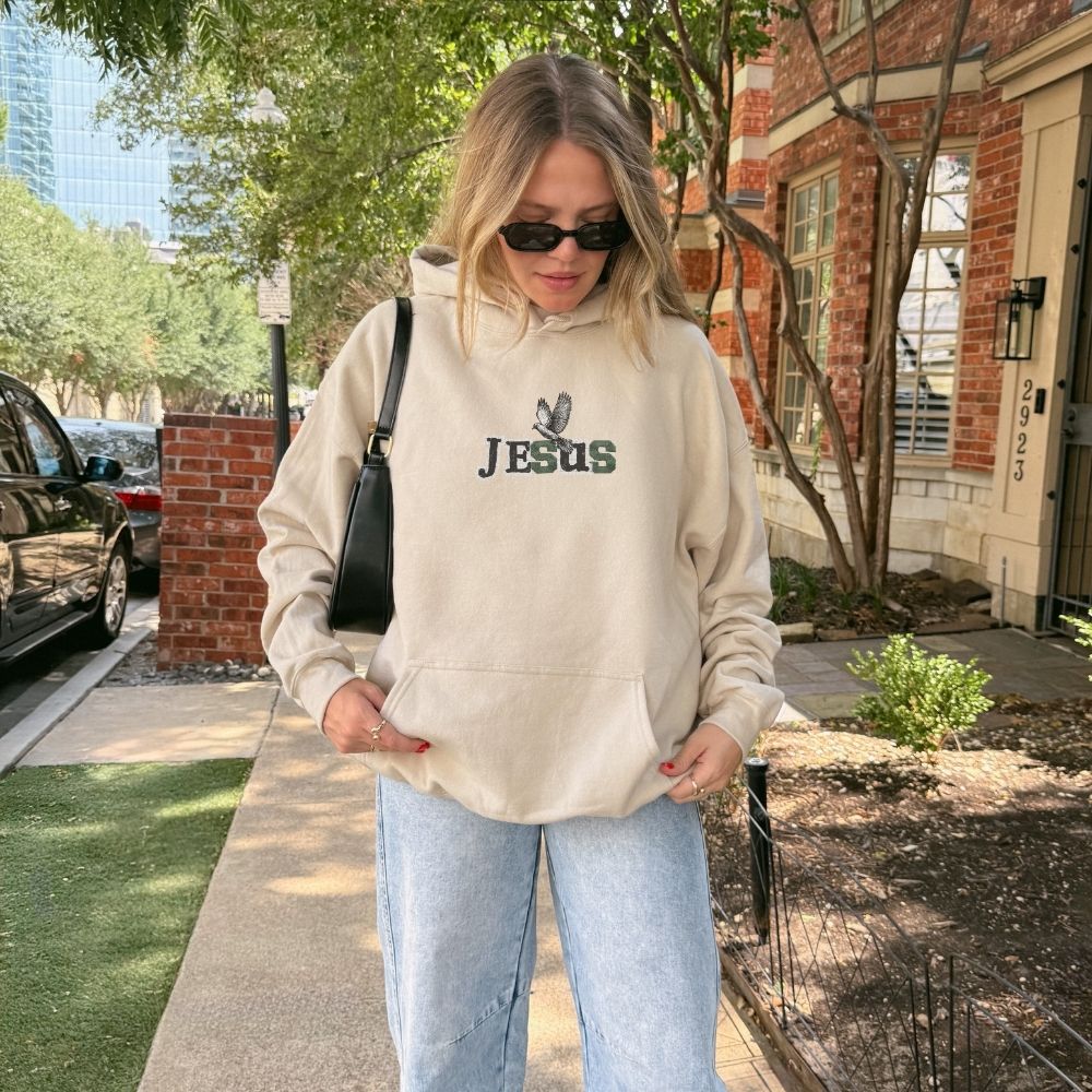 Person wearing a beige hoodie with 'Jesus' printed on it, standing outdoors.