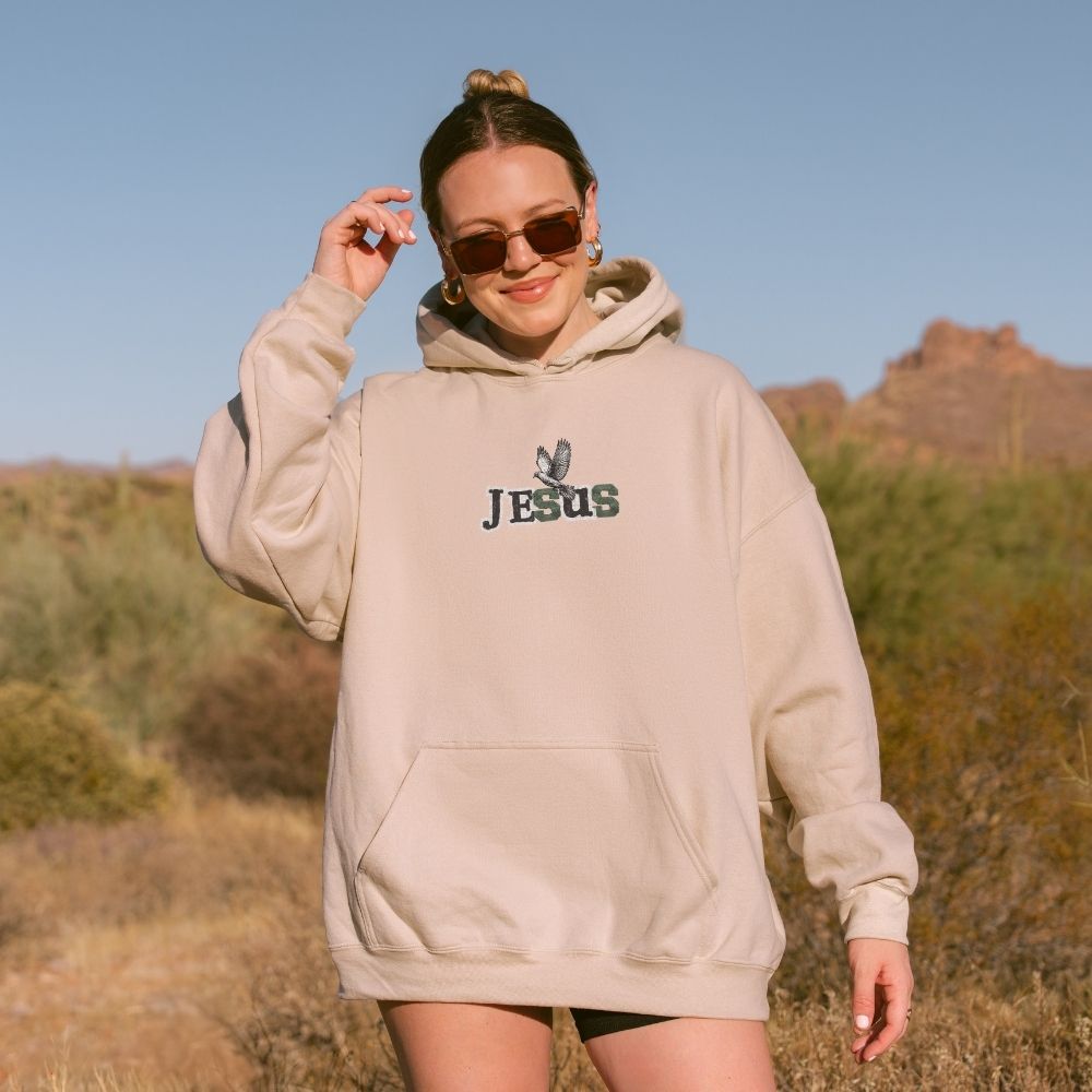 Person wearing a beige hoodie with 'JESUS' printed on it, standing in a desert landscape.