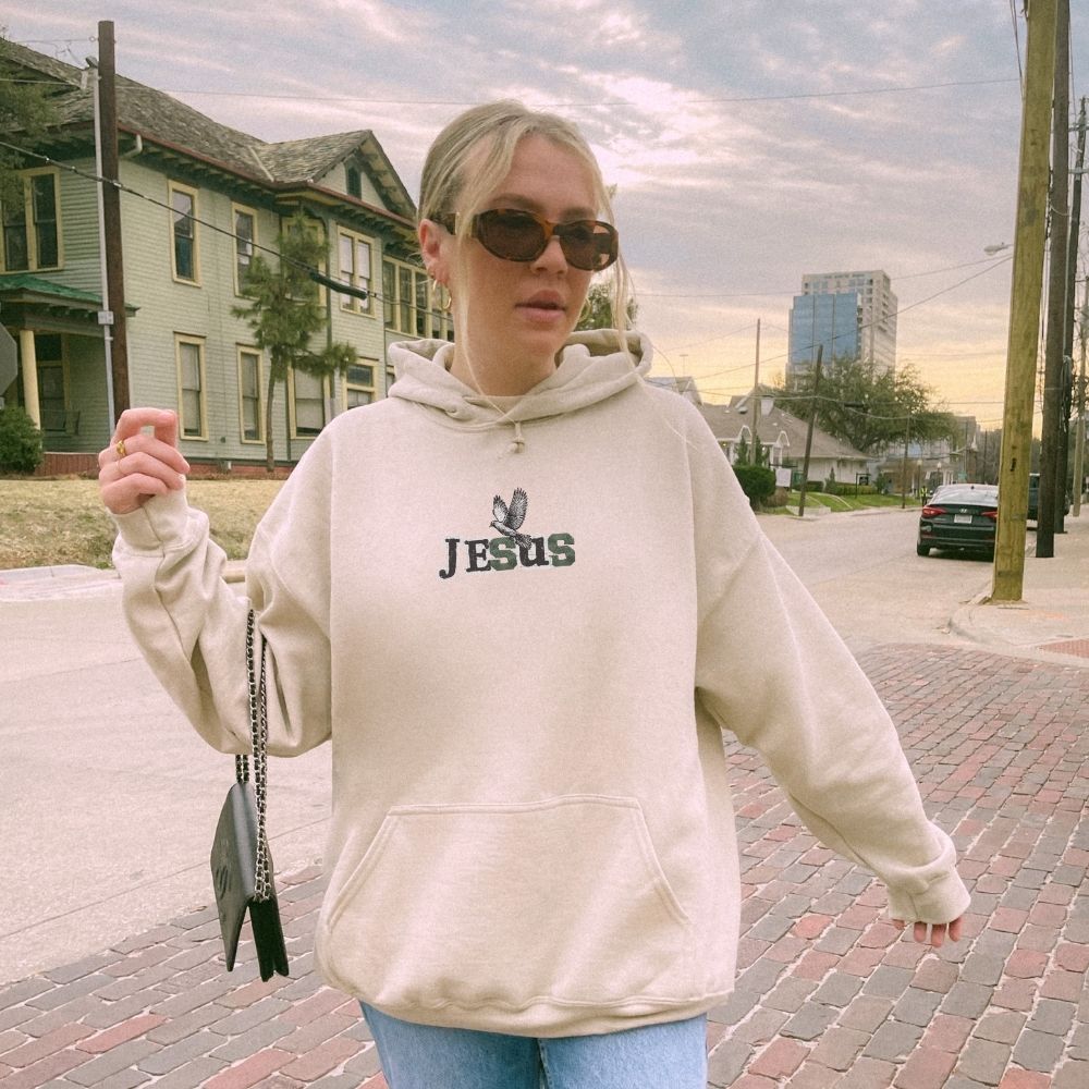 Person wearing a hoodie with 'Jesus' printed on it, standing on a street.