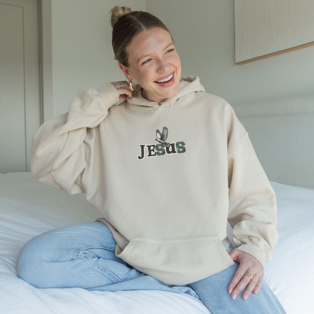 Person wearing a beige hoodie with 'Jesus' printed on it, sitting on a bed.
