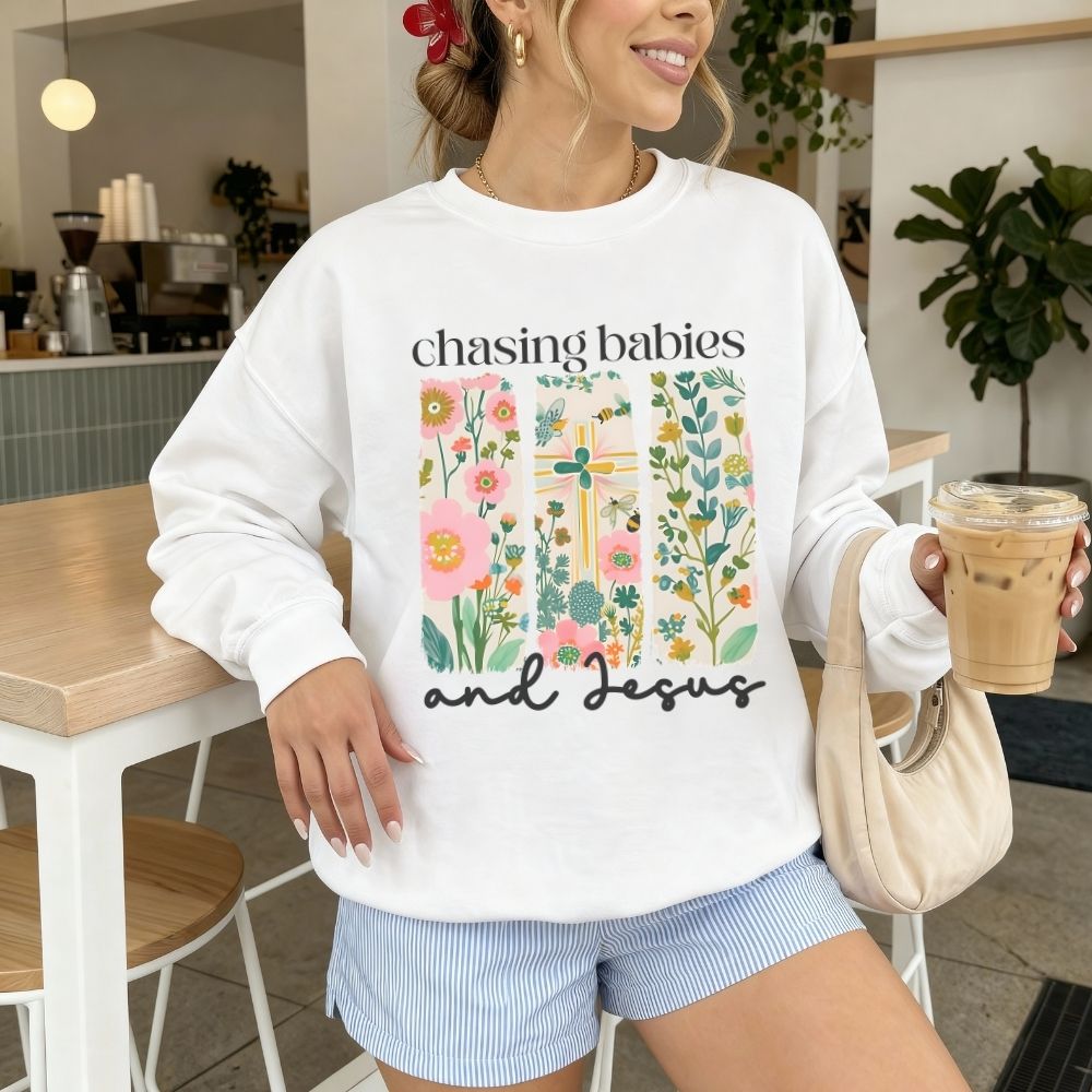 Person wearing a white sweatshirt with floral design and text, holding a coffee cup in a casual setting.