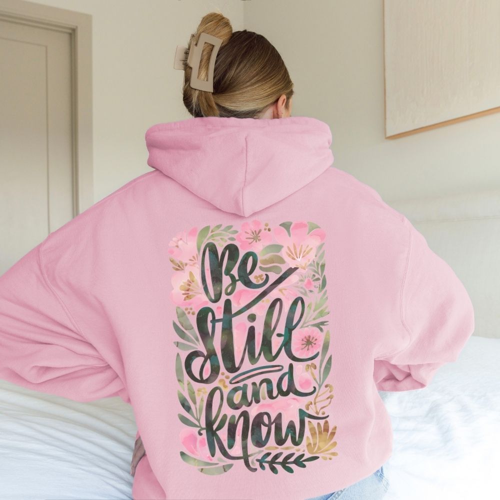 A person wearing a pink Christian hoodie with floral “Be Still and Know” design.