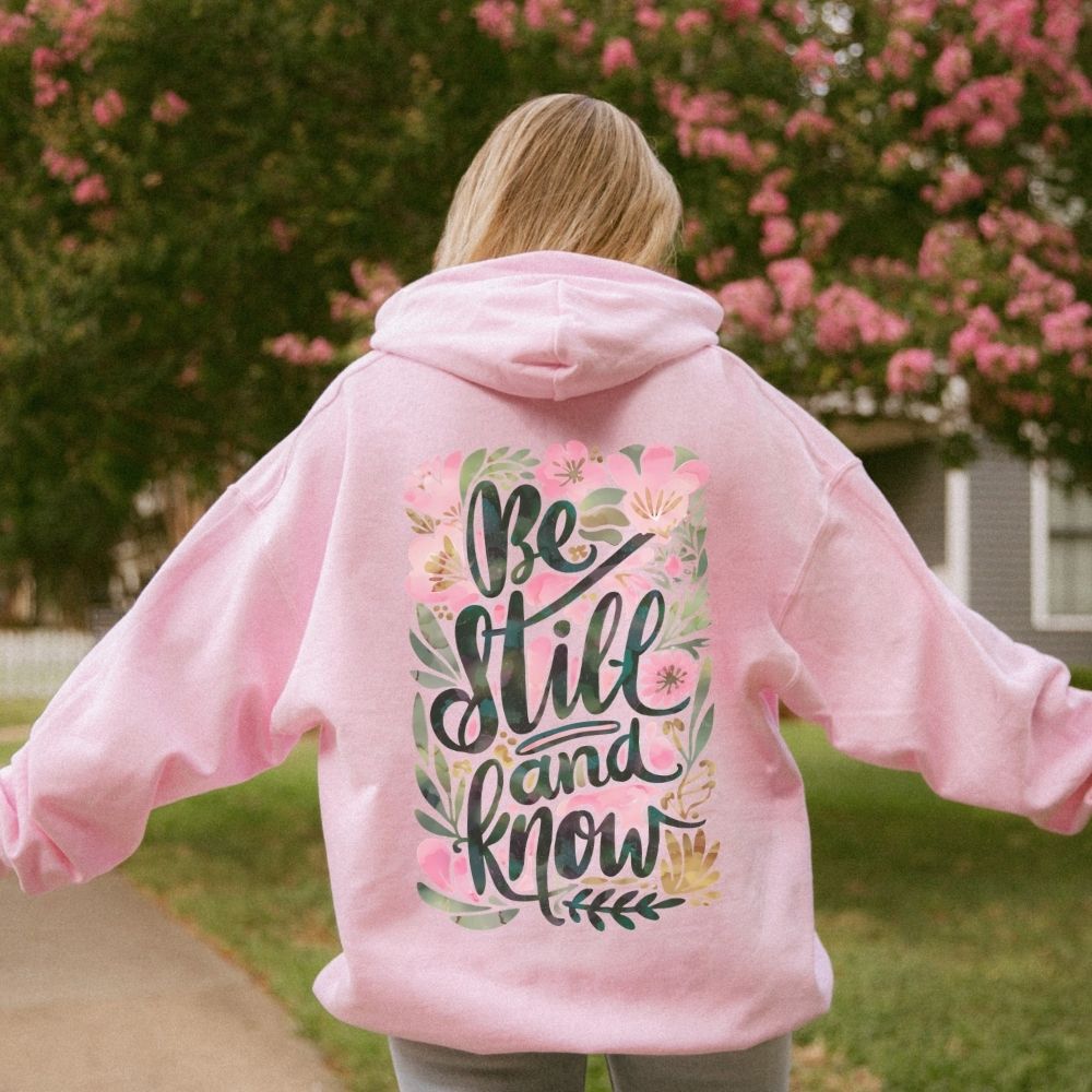 A person wearing a pink Christian hoodie with floral “Be Still and Know” design.