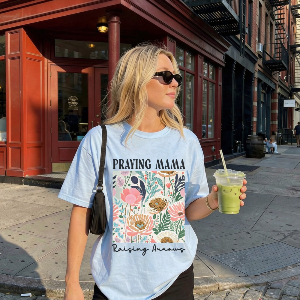 Woman wearing a 'Praying Mama' t-shirt with floral design, holding a drink on a city street.