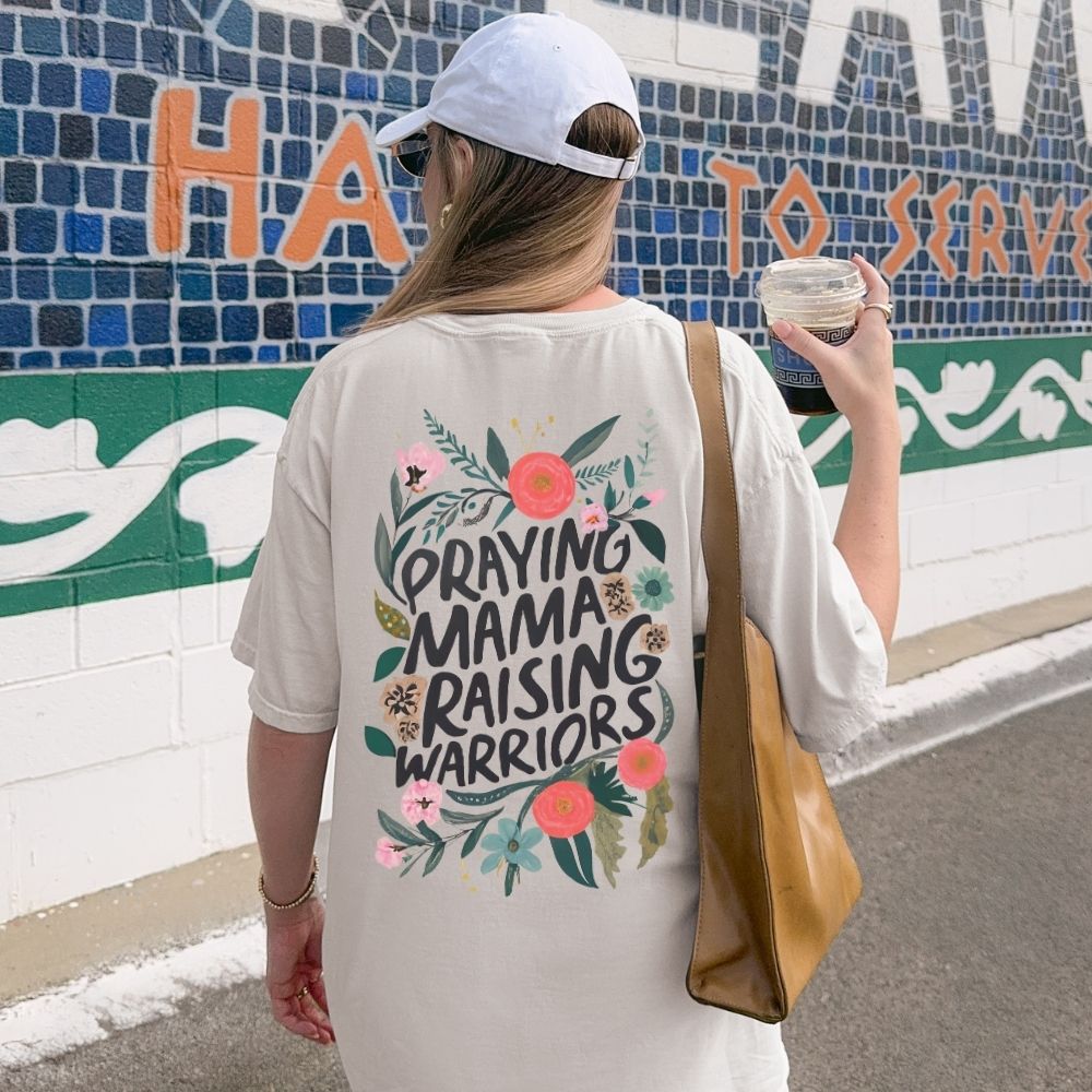 Woman wearing a t-shirt with 'Praying Mama Raising Warriors' design, holding a drink, standing in front of a mosaic wall.