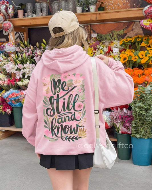 Young woman wearing a pink Christian hoodie with floral “Be Still and Know” design while shopping at a flower market.