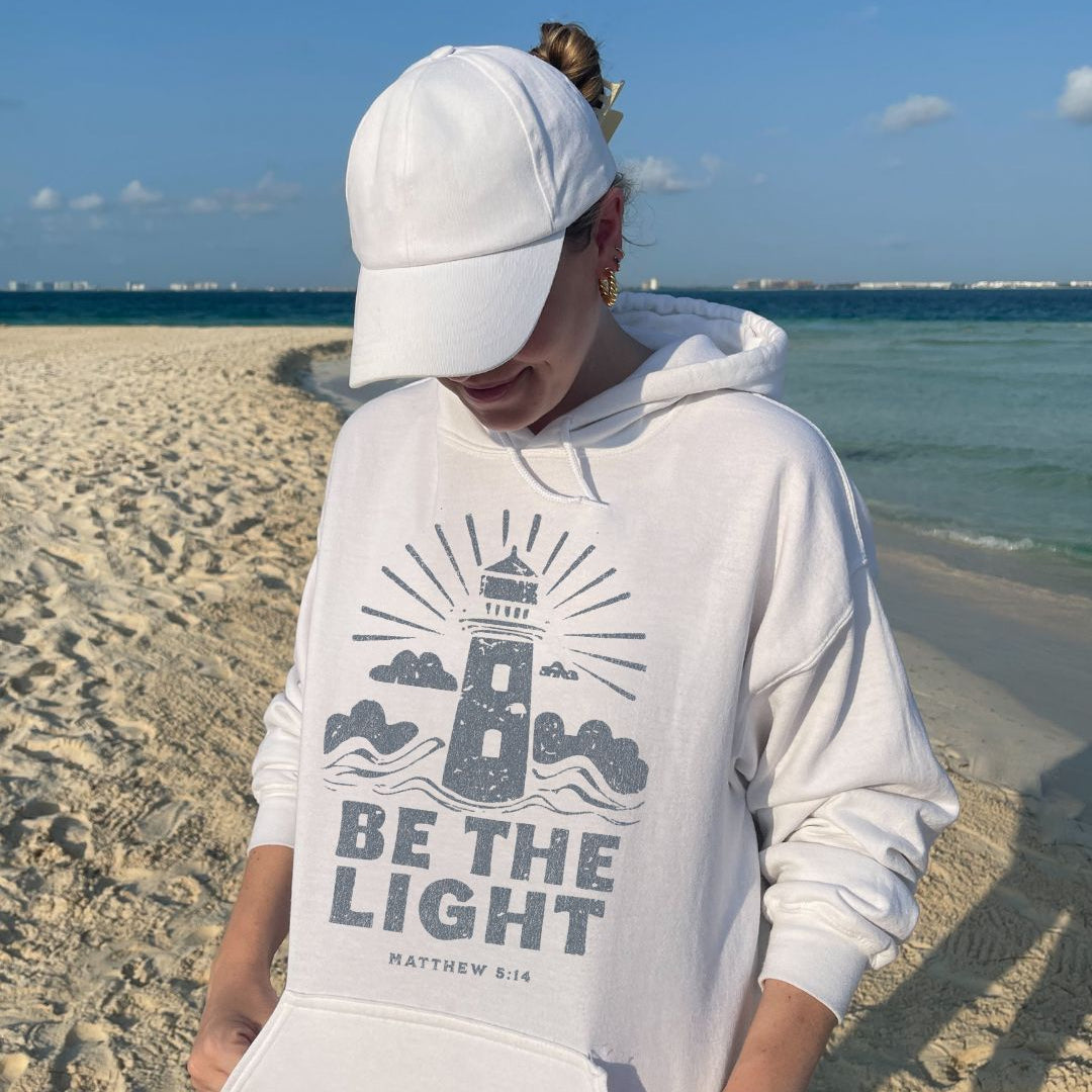 Woman wearing white Be The Light Christian hoodie on beach