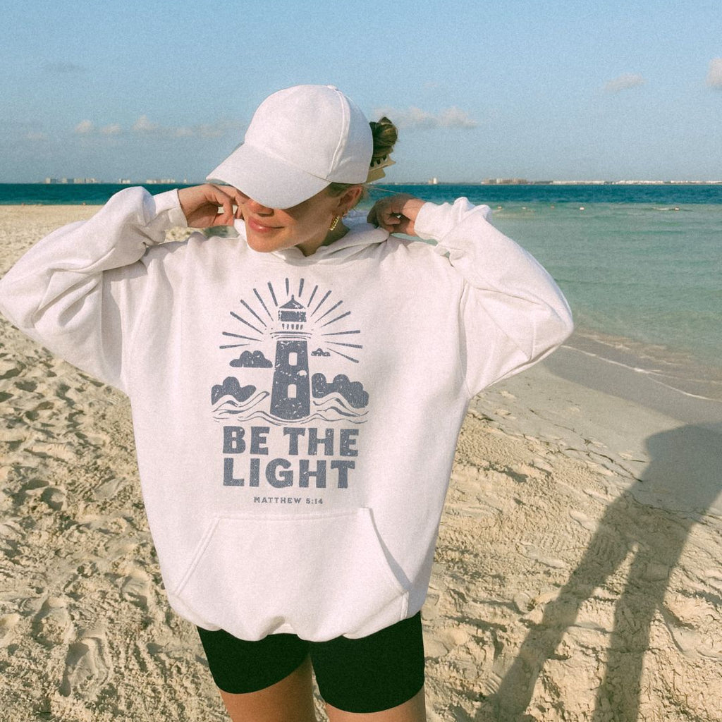 Woman wearing white Be The Light Christian hoodie on beach