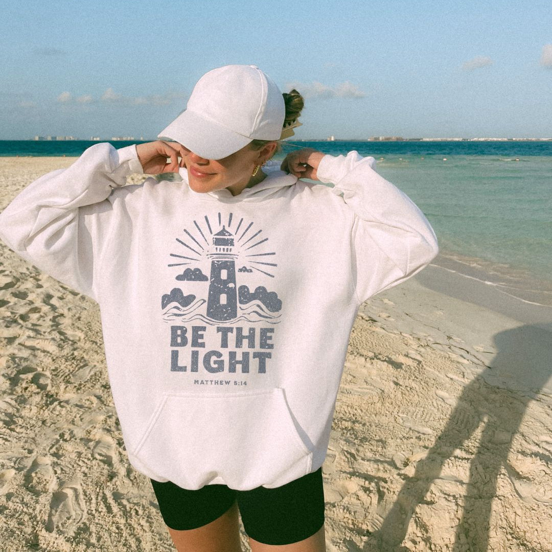 Woman wearing white Be The Light Christian hoodie on beach