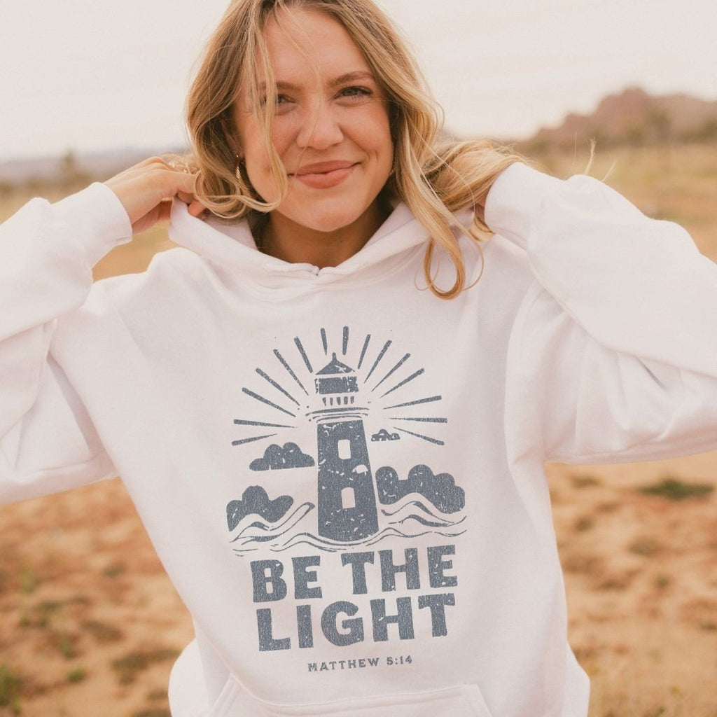Woman wearing a white hoodie with a lighthouse design and 'Be the Light' text in a desert setting.