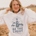 Woman wearing a white hoodie with a lighthouse design and 'Be the Light' text in a desert setting.