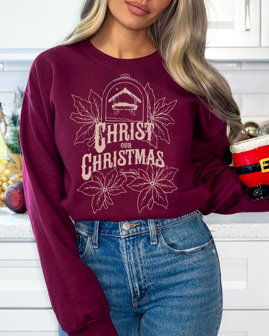 Woman wearing burgundy Christ Our Christmas sweater with manger and poinsettia design.