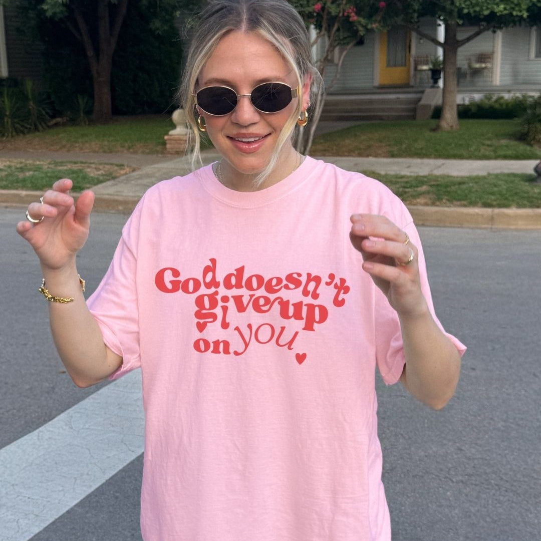Woman wearing a pink Christian t-shirt with bold red “God Doesn’t Give Up on You” design 